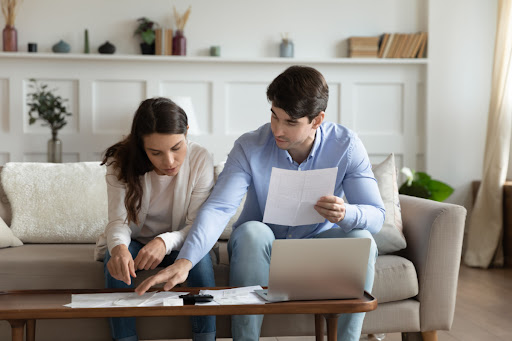 A man and woman sitting on a couch and looking at documents and a laptop.