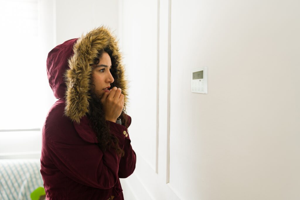 Woman in winter coat looking at thermostat while warming up her hands because her house is cold due to a broken heater.