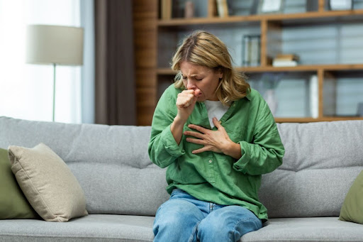 Woman sitting on her living room couch holding her chest while coughing into hand due to bad indoor air quality.