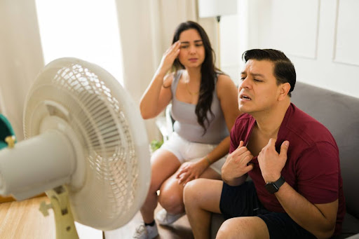 A man and a woman sitting in front of a fan in a hot house.